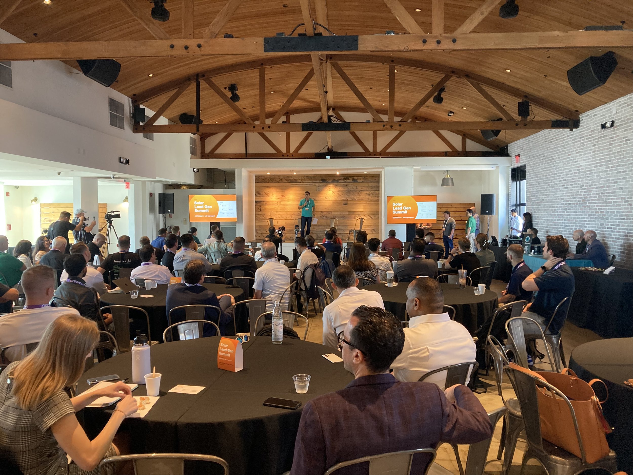 A conference room filled with attendees watching a speaker on stage during a Solar Lead Gen Summit presentation. The room has a high wooden ceiling and brick walls, with large TV screens displaying the event title.