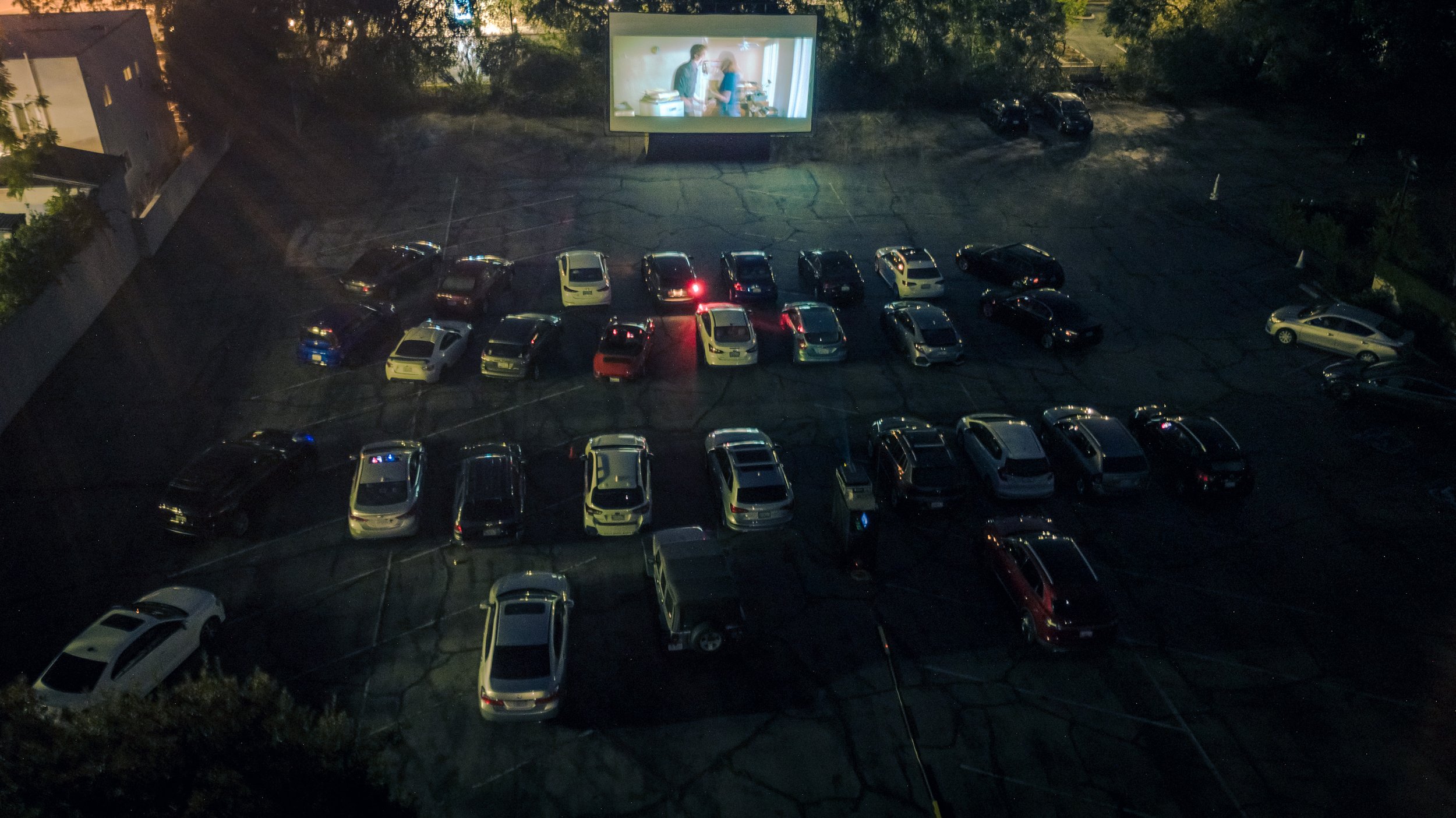 A drive-in movie theater at night with a large screen showing a scene with two people, and parked cars facing the screen.