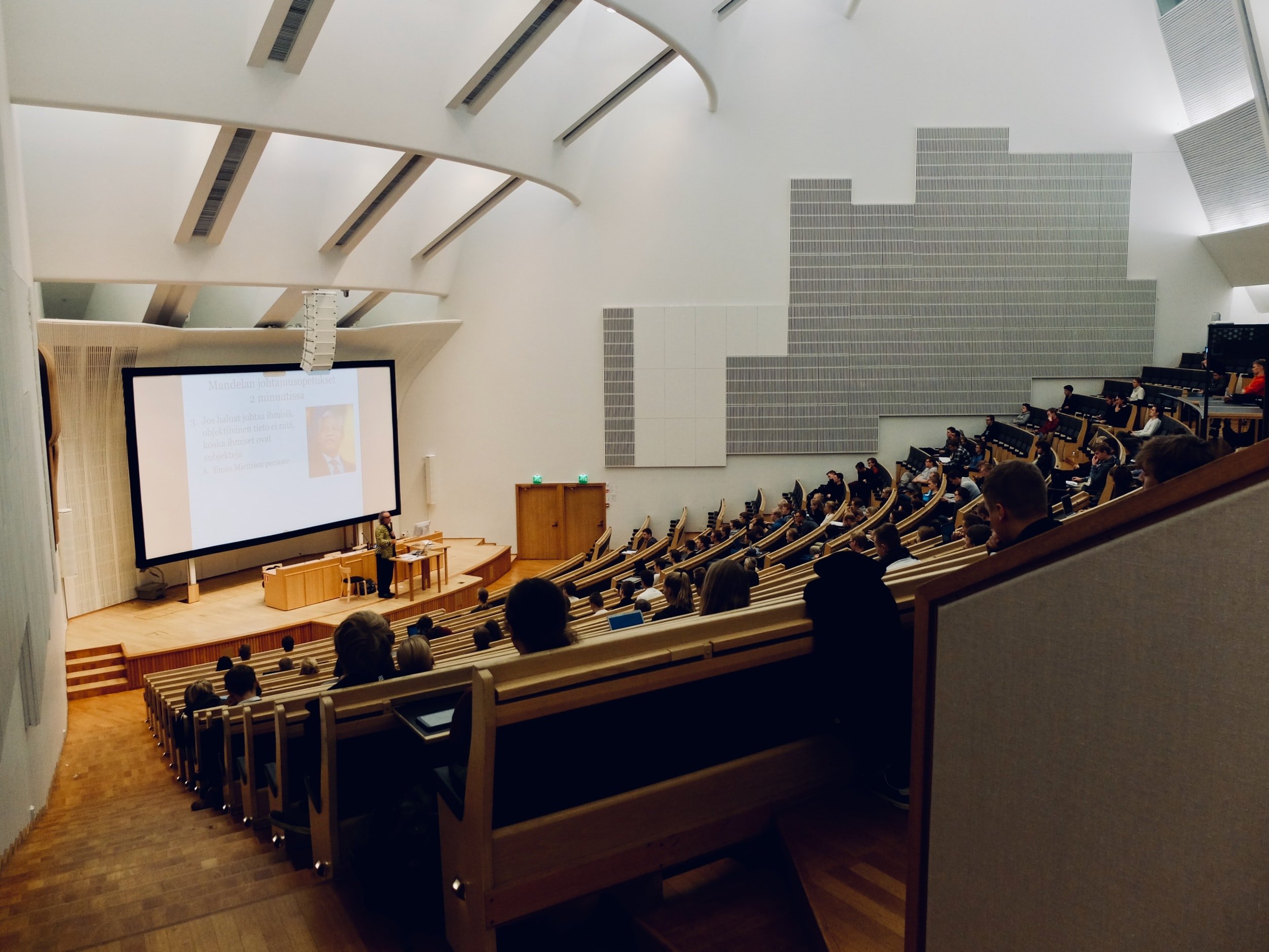 A lecture hall with curved seating filled with people, a speaker at the front, and a large screen displaying a presentation.